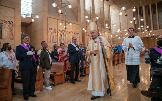 A light-skinned man in golden vestments and a mitre with a crozier walks down a cathedral aisle