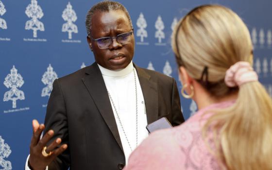 A Black man wearing glasses and a white collar talks to a blond woman who holds a phone towards him