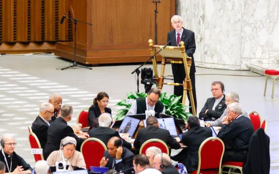 man in a dark suit and red tie stands at lectern and speaks to crowd, A full table of people sits in front of him