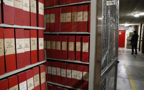 Red binders or upright boxes line metal shelves in a basement. Some have dates on the spines.