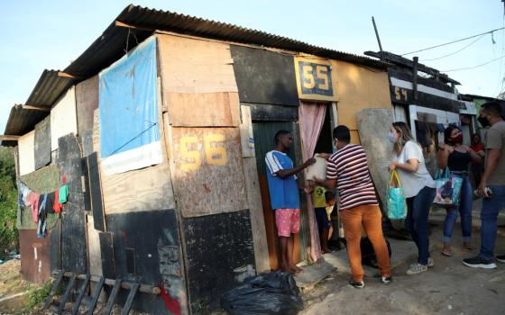 Masked people hold bags as they stand outside a house structure made of scrap wood and metal