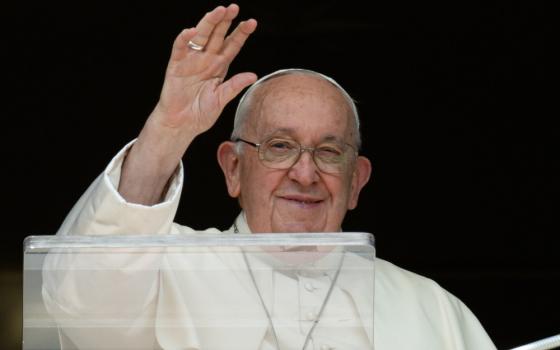 Pope Francis smiles and raises his white hand as he stands behind a clear lectern