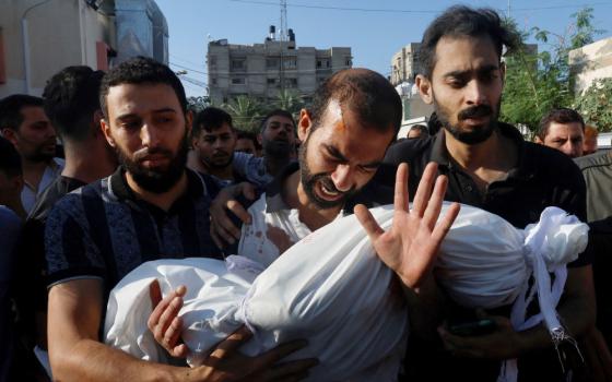 A man hangs his head and weeps, holding a body wrapped in a white sheet, while two men on either side of him hold him up