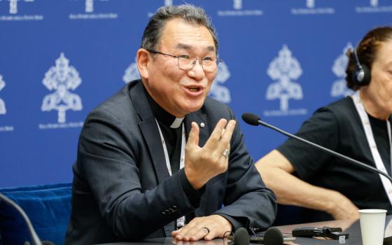 A smiling East Asian man wearing glasses and a clerical shirt and jacket speaks into a microphone