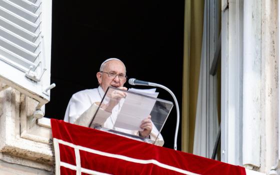 Pope Francis stands in his apartment window and holds a piece of paper above his clear lectern as he speaks into a microphone