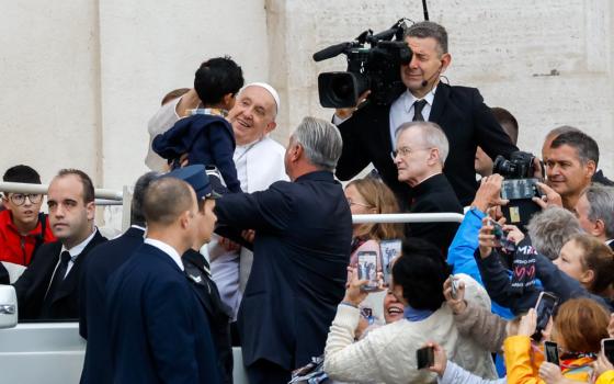 Pope Francis smiles at a child who is held up to him while he is riding in the popemobile. The crowd and a TV cameraman are visible around him.