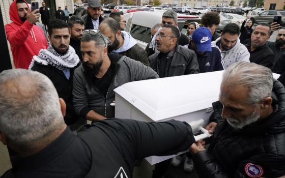 A group of brown men surround and hold an small white casket in a parking lot