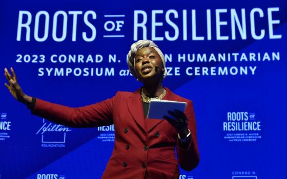 Woman holds papers and gestures with one arm. Behind her is written, "Roots of Reliliance."