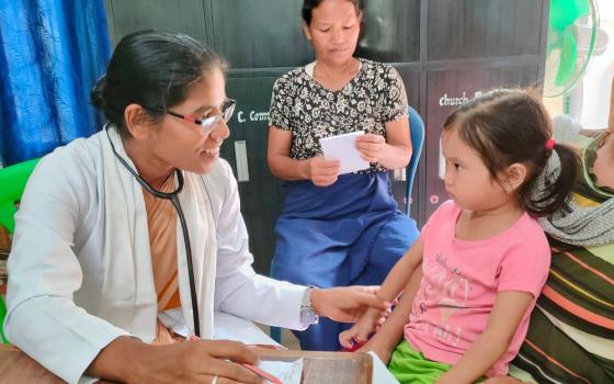 Woman wearing doctor's jacket and with stethoscope talks to little girl and touches her arm.