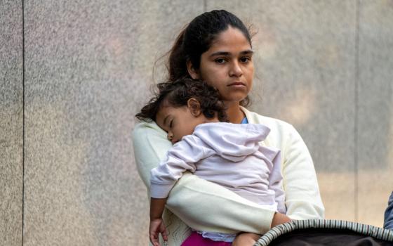 A young brown woman stands and holds a sleeping toddler. The woman looks serious.