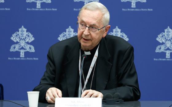 An older white man wearing glasses and a clerical collar speaks from behind a table and in front of a blue background with emblems 