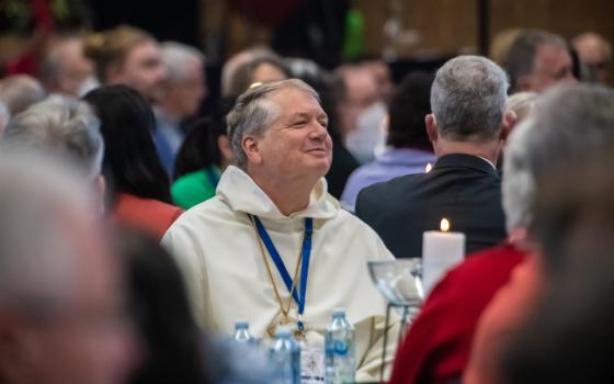 An older white man wearing a white robe and pectoral cross sits in a crowded room