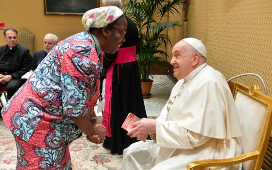 A Black woman wearing a colorful outfit and headscarf leans down to talk to Pope Francis, who is seated