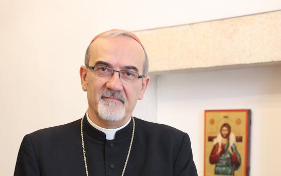 A white man wearing a cassock and a pectoral cross stands and looks at the camera