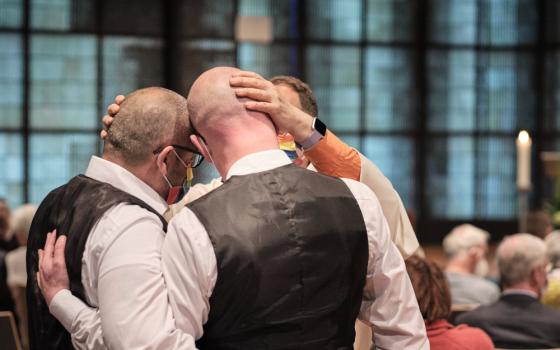 Two men in formal clothes stand close, heads touching, as a priest places his hands on both their heads