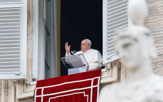 Pope Francis raises his right hand from the window of his apartment. A statue is in the foreground of the photo.