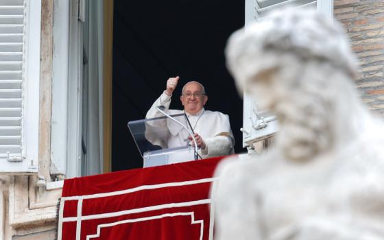 Pope Francis raises a thumbs up from his apartment window. A statue is visible in front of the shot of the window.