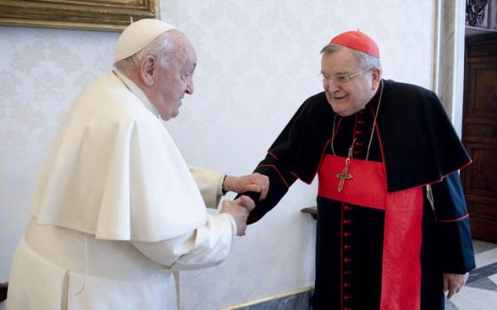 Pope Francis shakes the hand of an older white man wearing a red zucchetto and cardinal's cassock