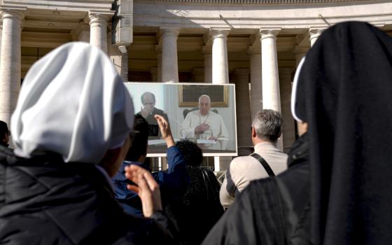 Pope Francis and a priest can be seen on a screen in front of a crowd with the backs of the heads of two religious sisters immediately in front of the camera