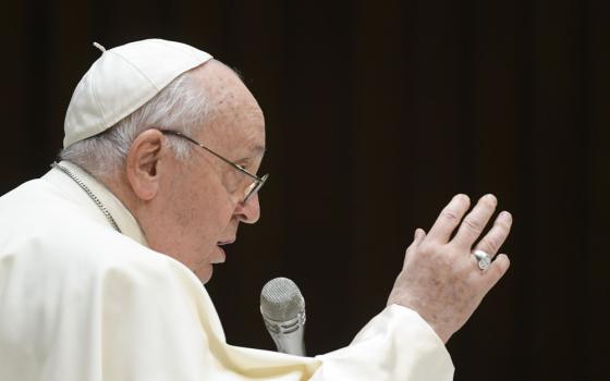 Pope Francis raises his right hand as he speaks into a microphone. He is against a black background.