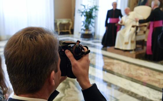 A white man raises a camera. Pope Francis and two bishops can be seen blurry in the background.