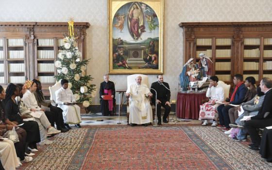 Pope Francis sits in a chair in a carpeted room with bookshelves. Attendees, including many young people of color, sit on the sides of the room.