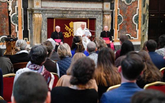 Pope Francis sits in front of a crowd, holding a piece of paper and speaking into a microphone