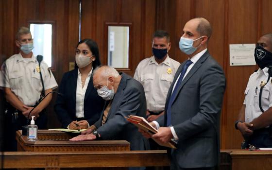 An older white man leans over behind a stand in a courtroom, where everyone is wearing masks