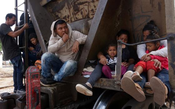 People sit on the side of a freight train car. Two women grip toddlers in their laps.