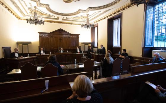 Many men sit at the front of a room below a crucifix with observers seated in chairs facing them