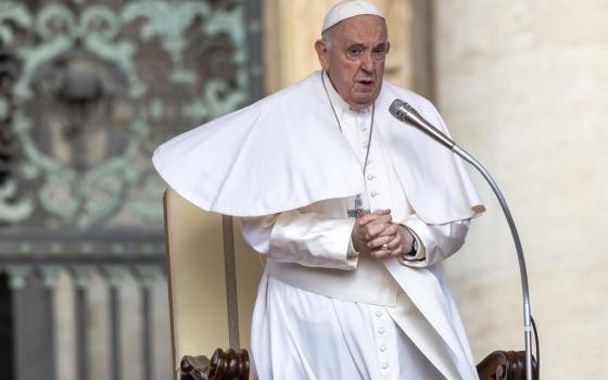 Pope Francis stands, hands folded, bracing against wind. 