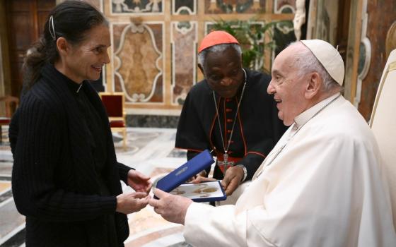 Pope Francis seated, accepts item from woman standing, cardinal bends to join.