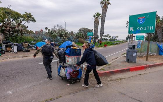 Two people push a cart filled with items across freeway toward encampment