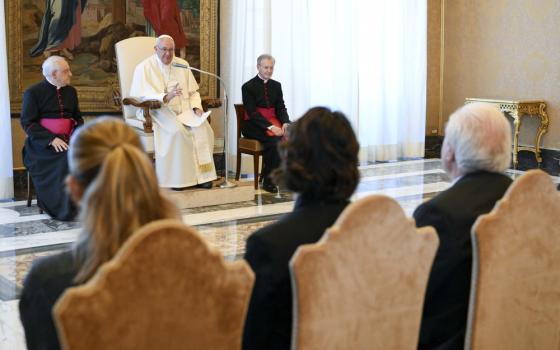 Pope Francis seated, flanked by Cardinals, people sit facing him, the back of their heads to the camera. 