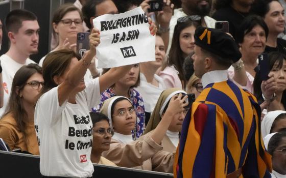 Woman wearing shirt reading "Stop blessing corridas" stares down Swiss Guard and holds up sign reading "Bullfighting is a sin."