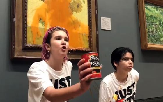 Two young people wearing matching t-shirts, stand in front of painting; one holds can of soup, whose contents are splattered across painting. 