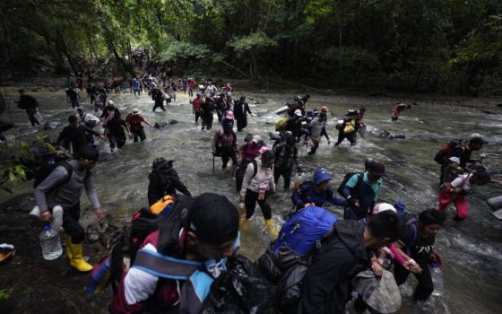 People wearing backpacks and other burdens walk through running water in jungle terrain. 