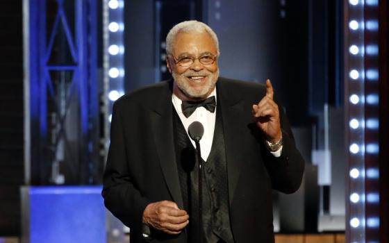 Jones, wearing formal wear, speaks from stage; hand raised in gesture, beaming a large smile.