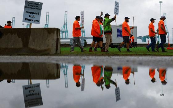 Picket line viewed from side, as workers wearing reflective orange gear and holding signs walk by; they are reflected in a pool of water that occupies the bottom half of the frame. 