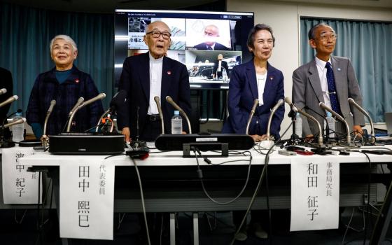 Four elderly people stand behind a table with microphones at press conference; behind is a screen.