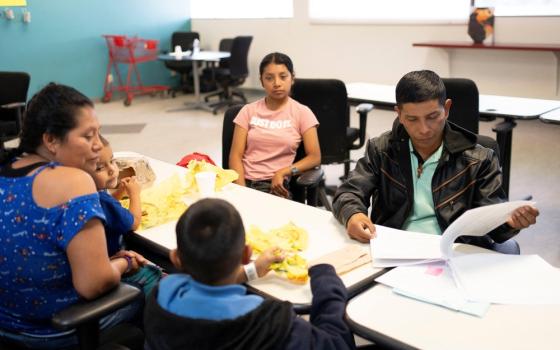 Family sits around table with trays of food in what looks like a classroom.