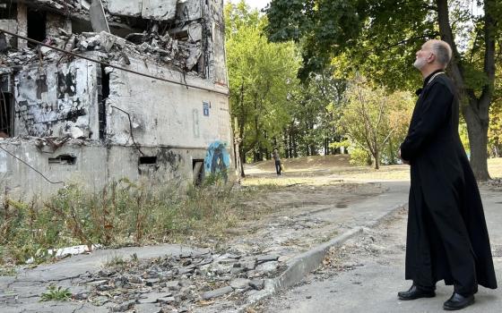 The archbishop wears plain black cassock, and stands back regarding ruins of building.