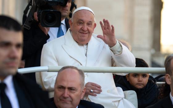 Francis seated, hand lifted in greeting and smiling.