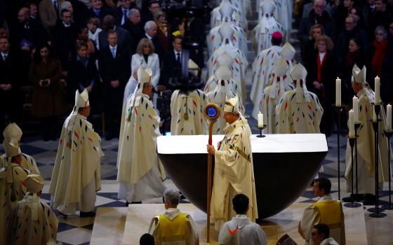 Archbishop, fully vested, pictured in profile holding crozier with massive blue stone; other bishops process, and seated in front of right row are France's presidential couple.