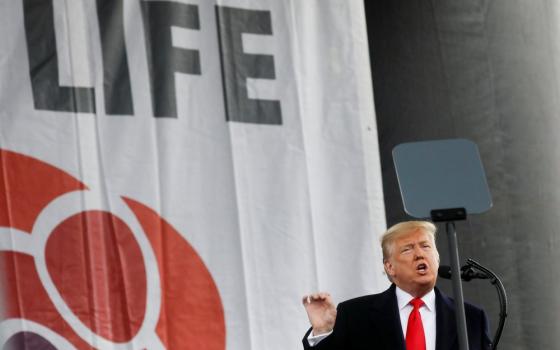 Trump stands at podium in front of large banner gesturing while talking.