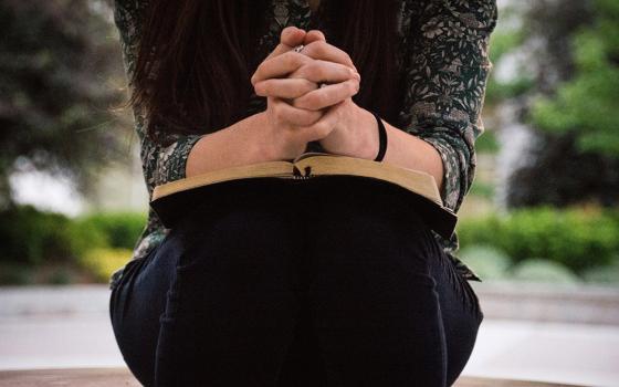 A woman sits outside with her hands folded on top of an open book, possibly a bible. (Unsplash/Olivia Snow)