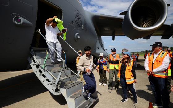 Two men step off of plane onto tarmac awaited by people wearing orange reflective vests.