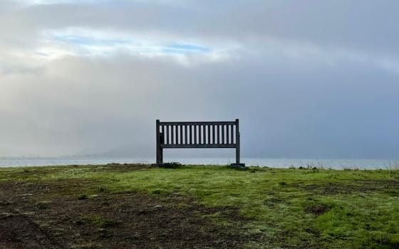A lone bench on patchy grass overlooking a cloudy sky with patches of sunshine shining through.
