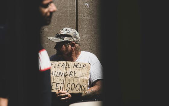 A person holds a sign saying "Please help, hungry" while another person walks by (Unsplash/Logan Weaver)