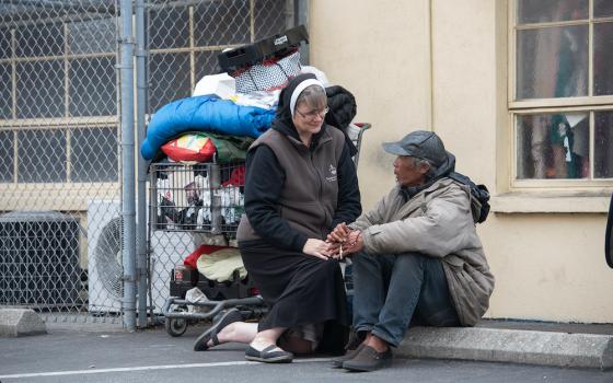 A habited nun kneeling to speak to an older man sitting on a curb next to a cart of his belongings